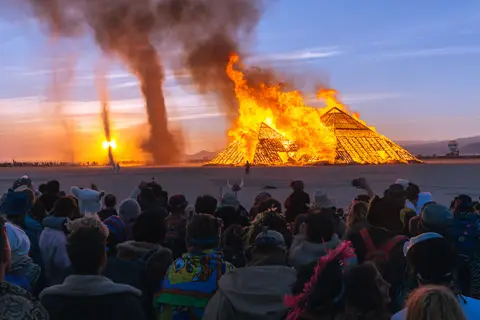 A massive wooden effigy burns brightly against a sunset sky, silhouetted by a crowd of festival-goers.