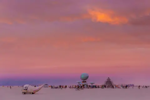 A surreal desert landscape unfolds under a vibrant pink and orange sunset sky, dotted with distant figures and unique art installations.