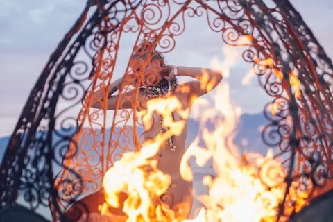 A person poses playfully within a fiery metal sculpture at dusk.
