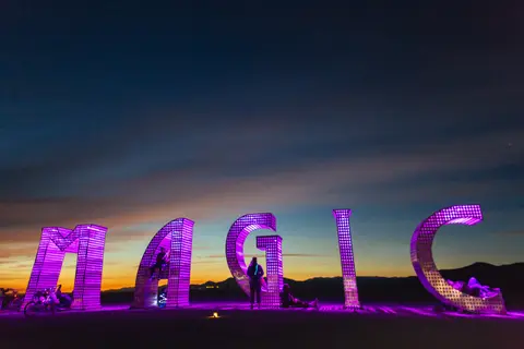 A lone figure stands silhouetted within the illuminated letters spelling 'MAGIC' against a dramatic twilight sky.