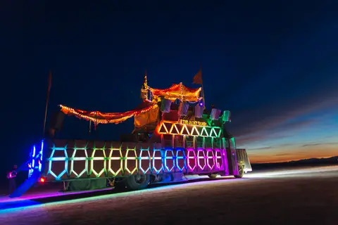 A brightly lit art car with colorful LED lights cruises across the playa at dusk during Burning Man.