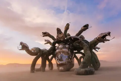 A striking sculpture resembling Medusa with multiple snake heads emerges from the dusty desert landscape under a dramatic sunset sky.