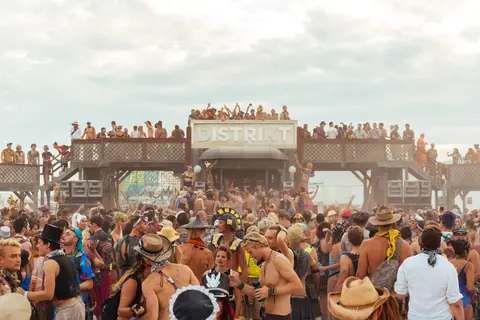 A massive crowd gathers in front of a large wooden structure with the word "DISTRICT" visible at a daytime outdoor festival.