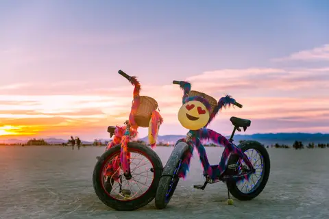 Two artfully decorated bicycles, one with a smiling emoji face, sit on a dusty plain under a vibrant sunset sky.