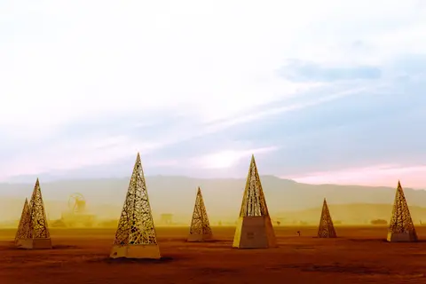 Geometric pyramid art installations dot a dusty landscape under a hazy, pastel sky.