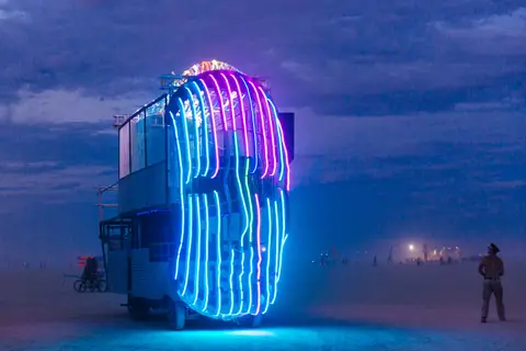 A large, illuminated art installation resembling a face glows with blue and pink neon lights against a twilight sky at Burning Man.