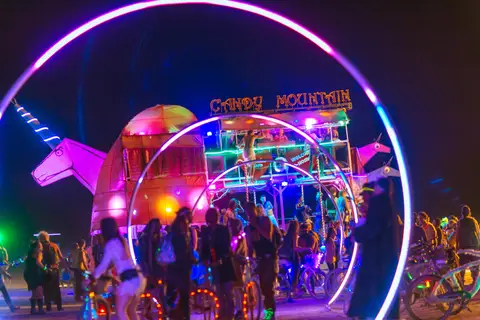 A vibrant night scene at Burning Man features a large, illuminated art installation with people gathered around.