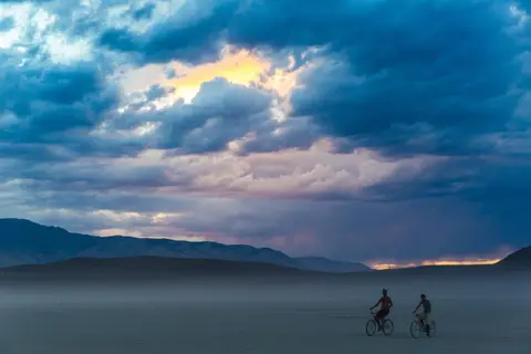 Two figures on bicycles ride across a vast, dusty plain under a dramatic, cloud-filled sky at sunset.