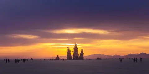 Silhouetted figures gather on a vast, flat plain as a dramatic sunset paints the sky in hues of orange, yellow, and purple behind a cluster