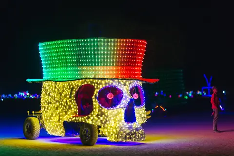 A brightly lit, skull-shaped art car adorned with a top hat covered in green and red lights cruises through the desert at night.