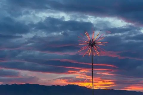 A lone, spiky art installation glows against a dramatic sunset sky at a desert festival.